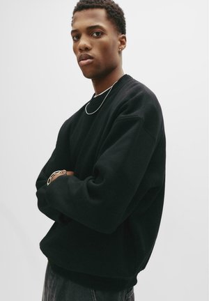Sort sweatshirt med rund halsudskæring og lange ærmer. Blødt stof, afslappet pasform og ribbede manchetter og kant. Model iført den mod en hvid baggrund.