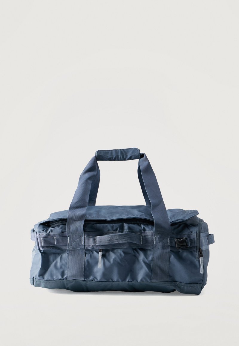 Σκούρο μπλε ορθογώνια τσάντα duffel με διπλές λαβές, πλαϊνές τσέπες και κλεισίματα με φερμουάρ σε απλό φωτεινό φόντο.