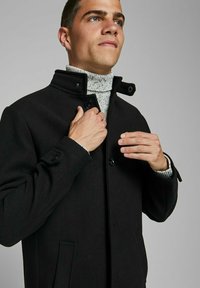 Melnas vilnas mēteles ar augstu apkakli, pogu aizdari un sānu kabatām. Vilkta virs strukturētā pelēkā turtlenecka džemperis.