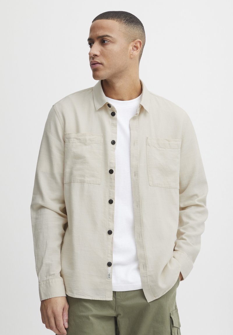 Camisa beige de botones con dos bolsillos en el pecho, botones negros y un corte relajado, usada sobre una camiseta blanca y pantalones verde olivo.