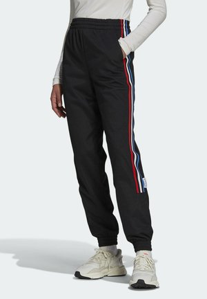 Personne portant un pantalon de survêtement noir avec des bandes latérales rouges, bleues et blanches, des baskets blanches, et un haut gris clair à manches longues.
