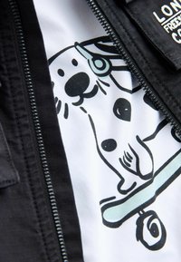 Črni suknjič z zadrgo, pod njim pa bela majica s prikupno risanko psa na rolki v črni in zeleno-modri barvi.