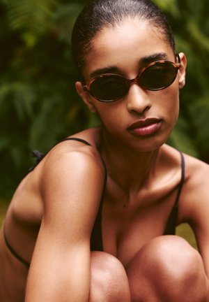 Ochelari de soare cu ramă din plastic cu model țestoasă, lentile ovale și brațe subțiri. Model cu păr scurt, purtând un top negru, stă într-un decor verde luxuriant.
