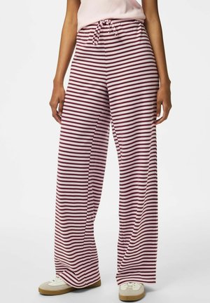 Mujer con pantalones de talle alto y pierna ancha con franjas horizontales granate y blancas y zapatillas blancas sobre un fondo liso.