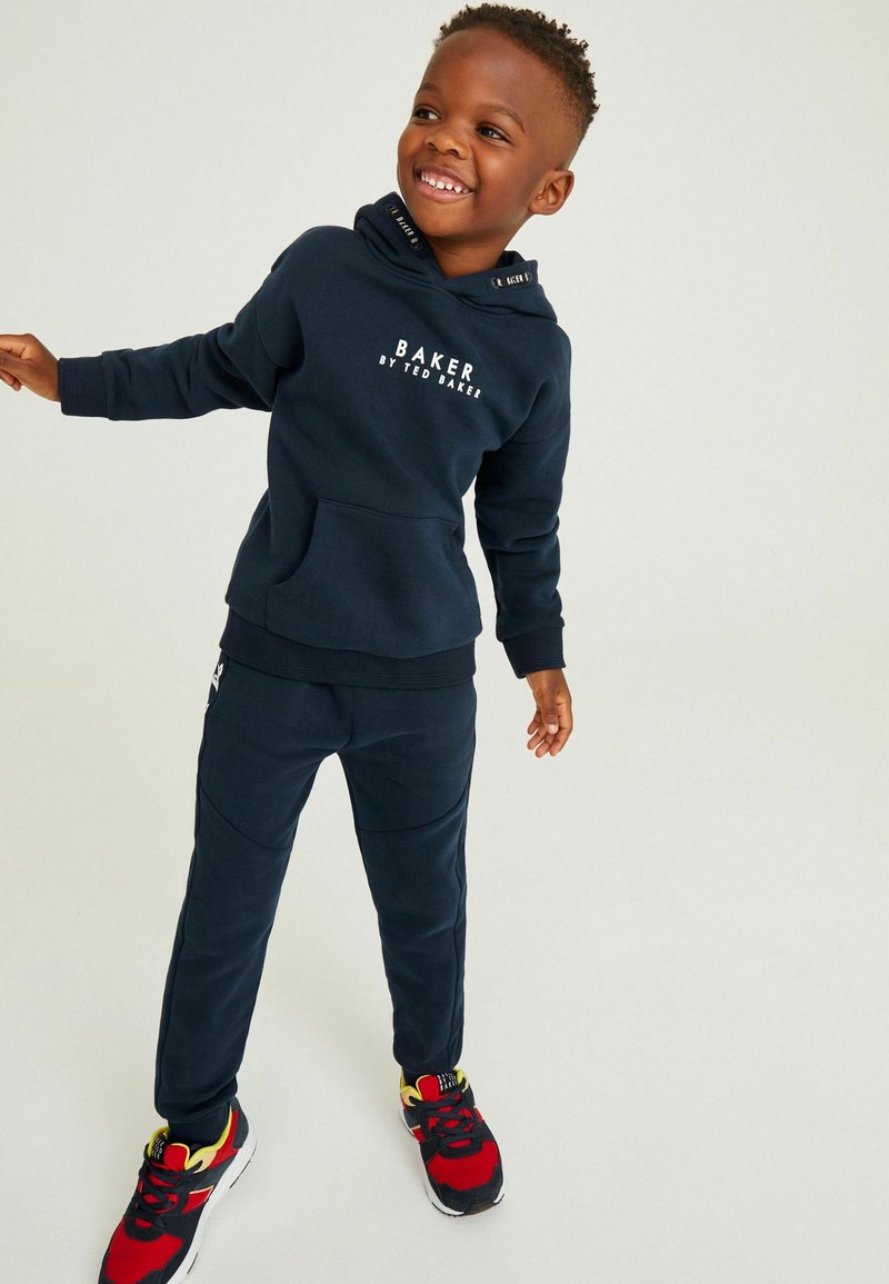 Set di felpa e pantaloni della tuta blu navy con tasca frontale, caratterizzato da testo del logo bianco. Completato con dettagli a contrasto su sneakers nere e rosse.
