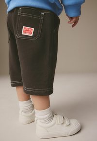 Barn med sorte shorts med en rød lap på baglommen, hvide sokker, hvide sneakers og en blå sweatshirt.