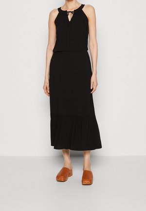 Femme portant une robe midi noire sans manches avec un col à nouer et des sabots marron fermés à talons carrés.