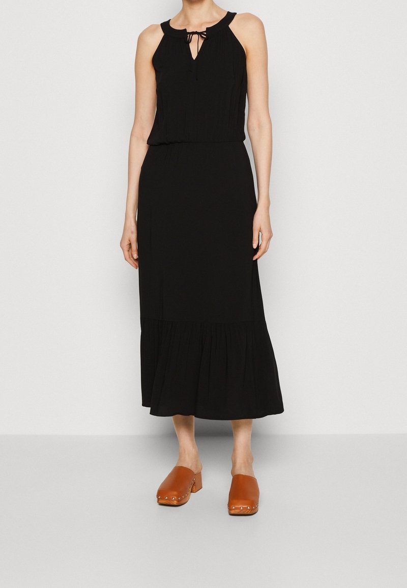 Femme portant une robe midi noire sans manches avec un col à nouer et des sabots marron fermés à talons carrés.