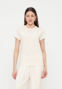 Γυναίκα με μακριά καστανά μαλλιά που φοράει χρυσούς κρίκους, κρεμ κοντομάνικη μπλούζα και κρεμ πλισέ παντελόνες με φαρδιά μπατζάκια, που στέκεται μπροστά σε λευκό φόντο.