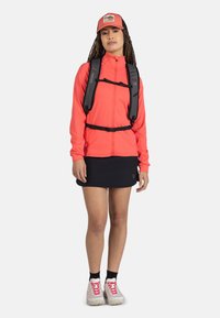 Lätt rosa zip-jacka, svart sportkjol, grå skor med rosa accenter, och en svart och röd keps. Ryggsäck med justerbara remmar.