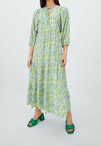 Frau in einem langen, lockeren Blumenkleid in Grün und Blau mit Dreiviertelärmeln und grünen Absatzsandalen vor einem schlichten Hintergrund.