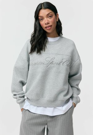 Kobieta z długimi ciemnymi włosami nosi jasnoszary sweter z wypukłym napisem "Universe Sport Club" oraz szare spodnie w prążki.