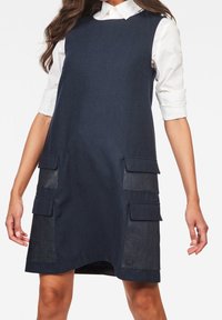 Mörkblå denimklänning med en avslappnad A-linjeform, två framfickor och en vit krag skjorta under.