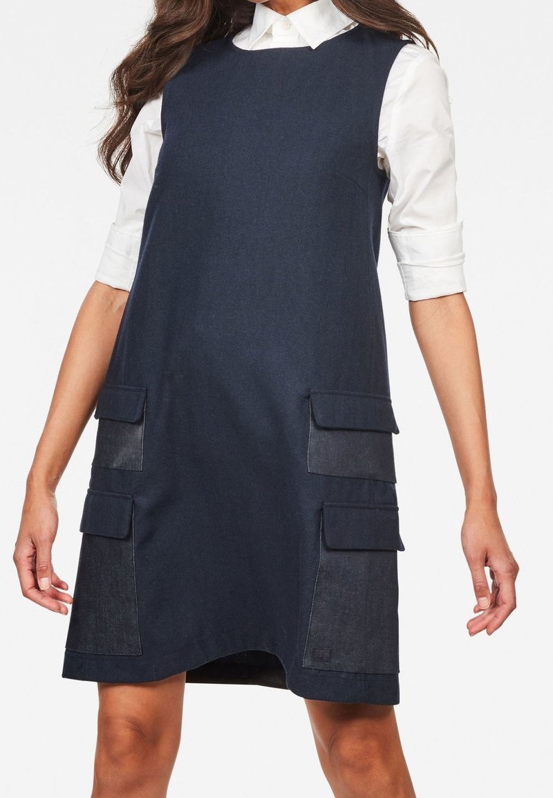 Mörkblå denimklänning med en avslappnad A-linjeform, två framfickor och en vit krag skjorta under.