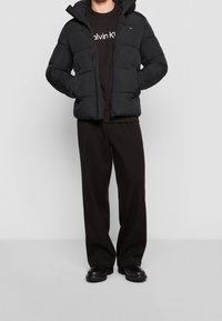 Czarna puchowa kurtka z kapturem, z poziomym przeszyciem i przednim zamkiem błyskawicznym, noszona na czarnej bluzie i dopasowanych spodniach.