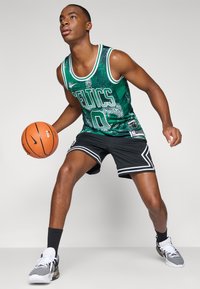 Groen basketbalshirt met witte accenten, korte broek met een zwarte basis en witte strepen, houdt een oranje basketbal vast, staand op een lichte achtergrond.