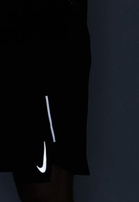 Persona vestida con pantalones cortos negros de Nike con logo y raya reflectantes, de pie contra un fondo oscuro.