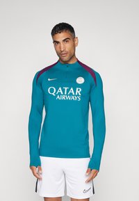 Teal urheilusetti korkealla kauluksella, maroonit olka-akselit, valkoinen "Qatar Airways" -logo ja pyöreä merkki. Yhdistetty valkoisten shortsien kanssa.