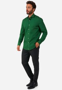 Camicia verde in cotone con una vestibilità slim, maniche lunghe e bottoni bianchi, abbinata a pantaloni scuri e scarpe nere.