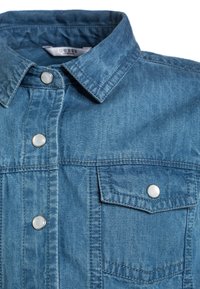 Světle modrá džínová košile s límcem, přední kapsou a stříbrnými patentkami. Vyznačuje se texturovanou tkaninou a viditelnými detaily šití.