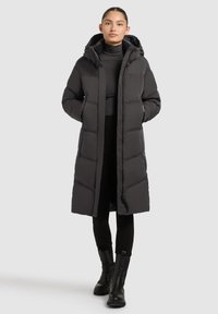 Sieviete stāv, valkājot garu melnu polsterētu mēteli ar kapuci, melnu augstu kakla džemperi, melnas bikses un melnas potīšu zābakus uz vienkārša fona.