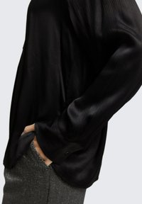 Blouse en satin noir avec des rayures texturées, coupe décontractée et manches longues. Les poignets présentent un détail de petit bouton. Portée avec un pantalon gris scintillant.