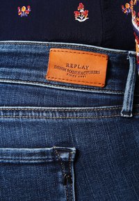 Los vaqueros de mezclilla presentan un parche de cuero marrón etiquetado "REPLAY DENIM MANUFACTURERS SINCE 1981", con costuras prominentes y un pequeño detalle del bolsillo.