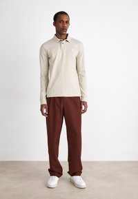 Beige langærmet polo skjorte med krave og diskret logo, parret med mørkerøde bukser og hvide sneakers på en neutral baggrund.