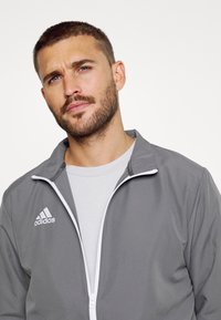 Graue Sportjacke mit weißen Akzenten und Reißverschluss, mit einem Adidas-Logo auf der Brust. Wird über einem hellgrauen T-Shirt getragen.