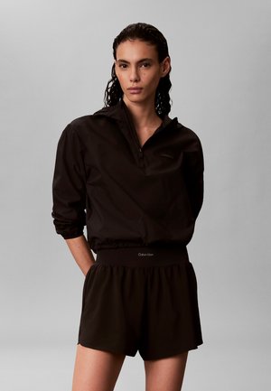 Nainen, jolla on märät hiukset ja joka pukeutuu mustaan pitkähihainen vetoketjulliseen paitaan sekä mustiin shortsseihin, joissa on joustava vyötärö, tasaisen harmaan taustan edessä.