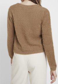 Brun fluffig tröja med långa ärmar, rund halsringning och texturerad finish; bärs med ljusa byxor som visar en avslappnad passform och avslappnad design.