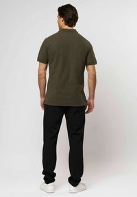 Muž stojící zády, oblečený v olivově zeleném polo tričku, černých kalhotách a bílých teniskách na neutrálním pozadí.