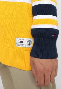 Pull-over en maille jaune avec rayures bleu marine et blanches. Présente des poignets côtelés et un patch logo avec texte à l'ourlet. Tissu texturé.