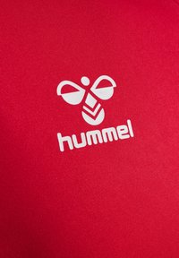 Tela roja con una textura suave presenta un logo blanco que se asemeja a una abeja y la palabra "hummel". Enfócate en el color vibrante y el diseño gráfico.