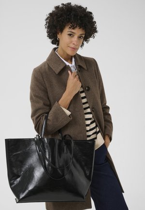 Schwarze, glänzende, künstliche Leder-Tasche mit zwei runden Griffen und geräumigem Design, kombiniert mit einem braunen Mantel über einem gestreiften Pullover.