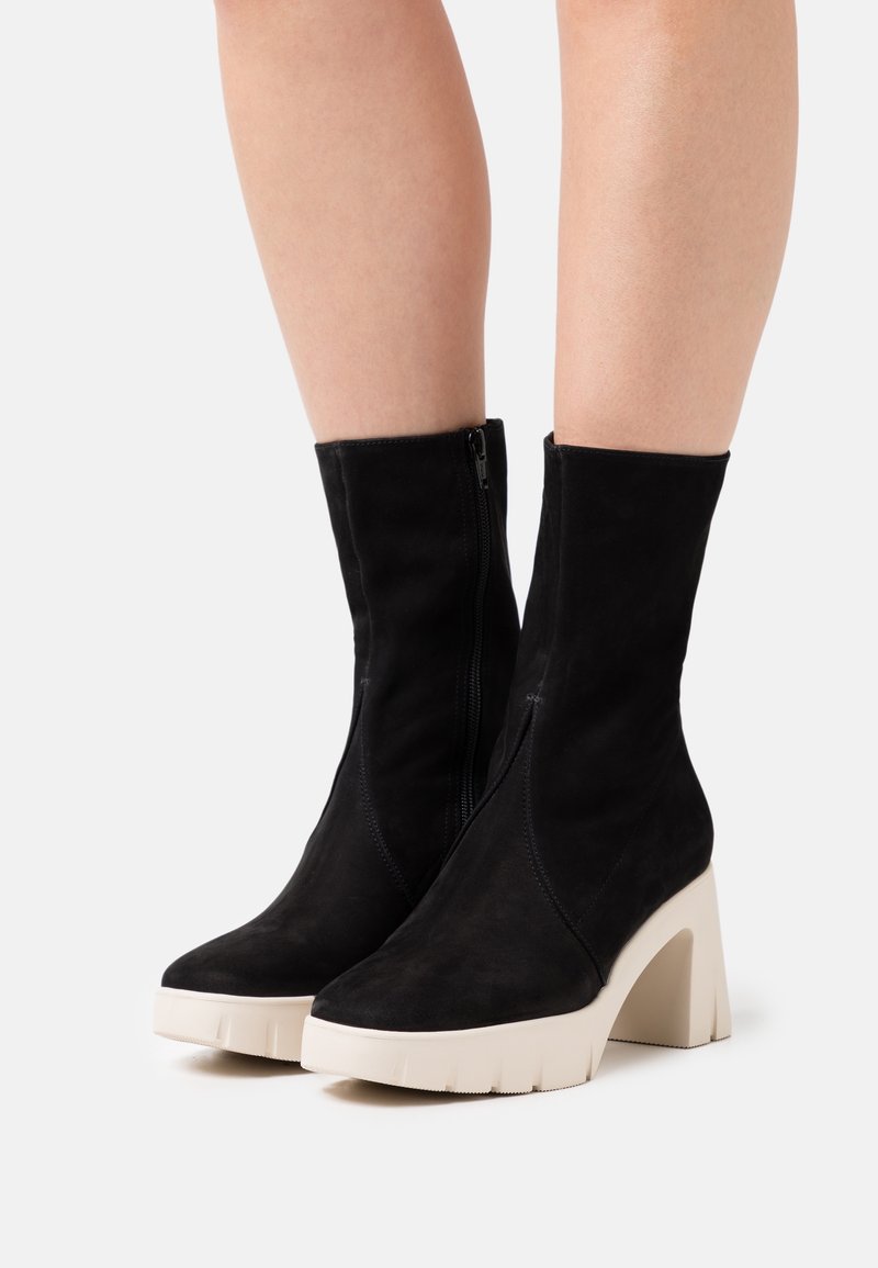 Bottes noires mi-mollet avec talons hauts blancs épais et fermetures éclair latérales portées sur des jambes nues sur un fond blanc.