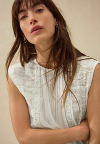 Blouse blanche avec des détails en dentelle intricats, un tissu plissé et un col haut. Texture lisse avec une coupe décontractée et un design sans manches.