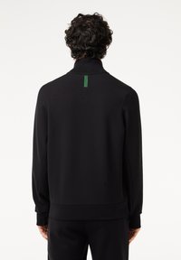 Sort sweatshirt med høj krave og lange ærmer, med et grønt mærke bagpå og en glat, blød tekstur.