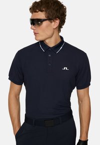 Marinblå pikétröja med två knappar, vita kragslinjer och en liten logotyp på bröstet. Bärs tillsammans med svarta solglasögon och ett bälte.