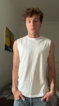 Young man with curly hair wearing a sleeveless white shirt and jeans, standing indoors with hands in pockets, neutral expression.