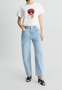Weißes T-Shirt mit einem bunten Grafikmotiv einer Frau auf der Vorderseite, kombiniert mit hellblauen, hochtaillierten, geraden Jeans und schwarzen, hochhackigen Sandalen.