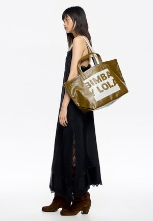 Olive green, błyszcząca torba tote z białym logo "BIMBA Y LOLA". Posiada mocne uchwyty i zorganizowany kształt. Nosi ją osoba w czarnej sukience.