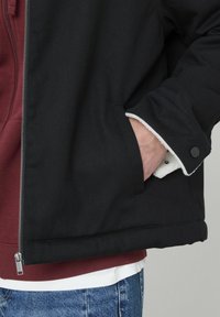 Veste zippée noire avec une texture lisse, dotée d'une capuche bordeaux en dessous, d'une doublure intérieure blanche et d'un poignet ajusté avec un bouton-pression noir.
