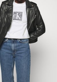 Musta nahkainen moottoripyöräjakku valkoisen, graafisen printin omaavan t-paidan päällä, yhdistettynä korkeavyötäröisiin sinisiin farkkuihin.