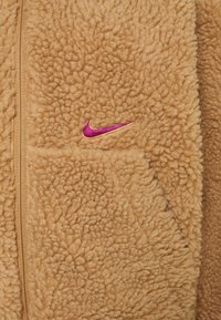 Nike fleecejacka i beige med en texturerad, fluffig yta, dragkedjestängning och en liten rosa logotypaccent på bröstet.