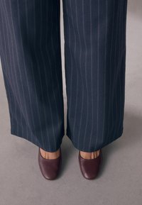 Vaasileveät tummansiniset housut, joissa on vaaleat raidoitukset, yhdistettyinä burgundinvärisiin nahkaisiin slip-on-kengiin. Materiaali on sileä ja malli rento.