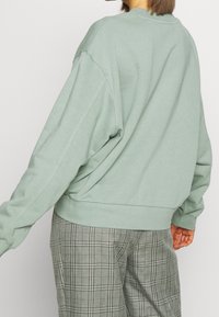 Person wearing a loose, light green sweatshirt paired with high-waist gray plaid trousers, standing against a plain light background.