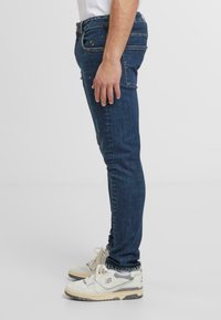 Siniset farkut slim fit -muodossa, joissa on keltaisia maalitippoja ja perinteinen viisi taskua -suunnittelu. Yhdistetty valkoisten ja harmaiden tennareiden kanssa.