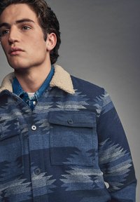 Navy patterned jacket with a beige shearling collar, featuring two front pockets and a subtle design in varying shades of blue.