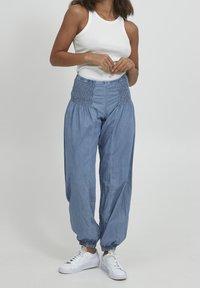 Mujer con una blusa sin mangas blanca, pantalones de tiro alto de smock en azul con puños elásticos, y zapatillas blancas, de pie contra un fondo liso.